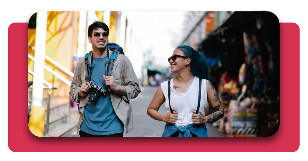 4 mejores apps para viajar que deberías tener en el celular 6 Dos personas con mochilas y una cámara fotográfica, sonríen mientras exploran un mercado popular urbano y ven las artesanías disponibles.